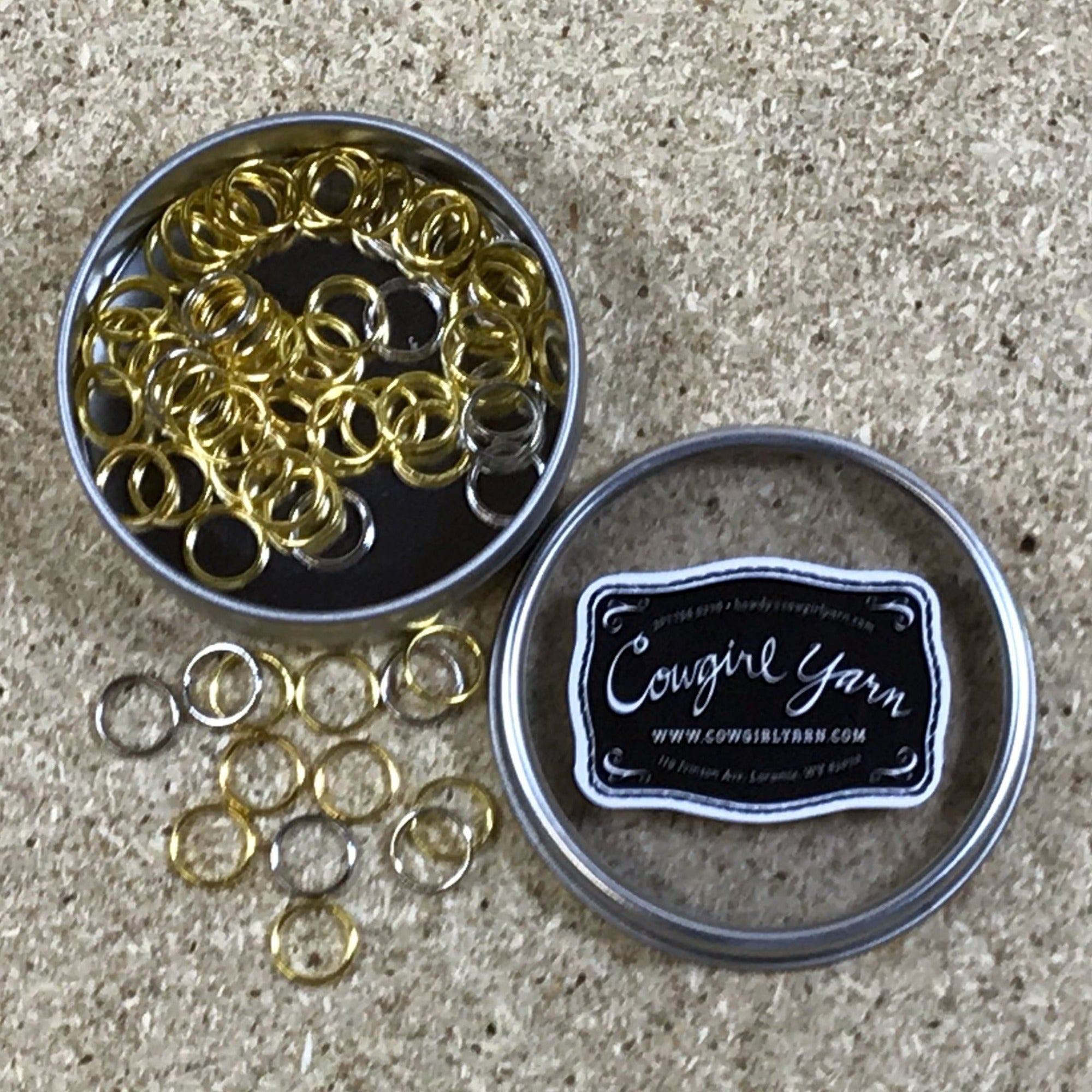 A Cowgirl Yarn branded metal tin filled with metal stitch markers in silver and bronze finishes
