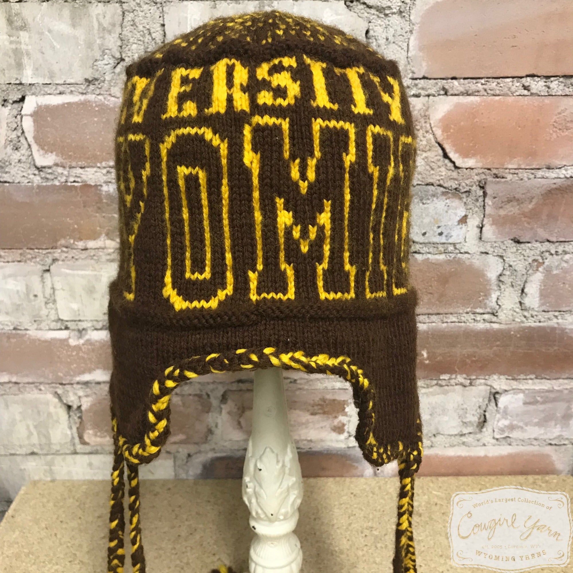 A brown knitted ski hat with the words University of Wyoming in gold styled against a brick wall.