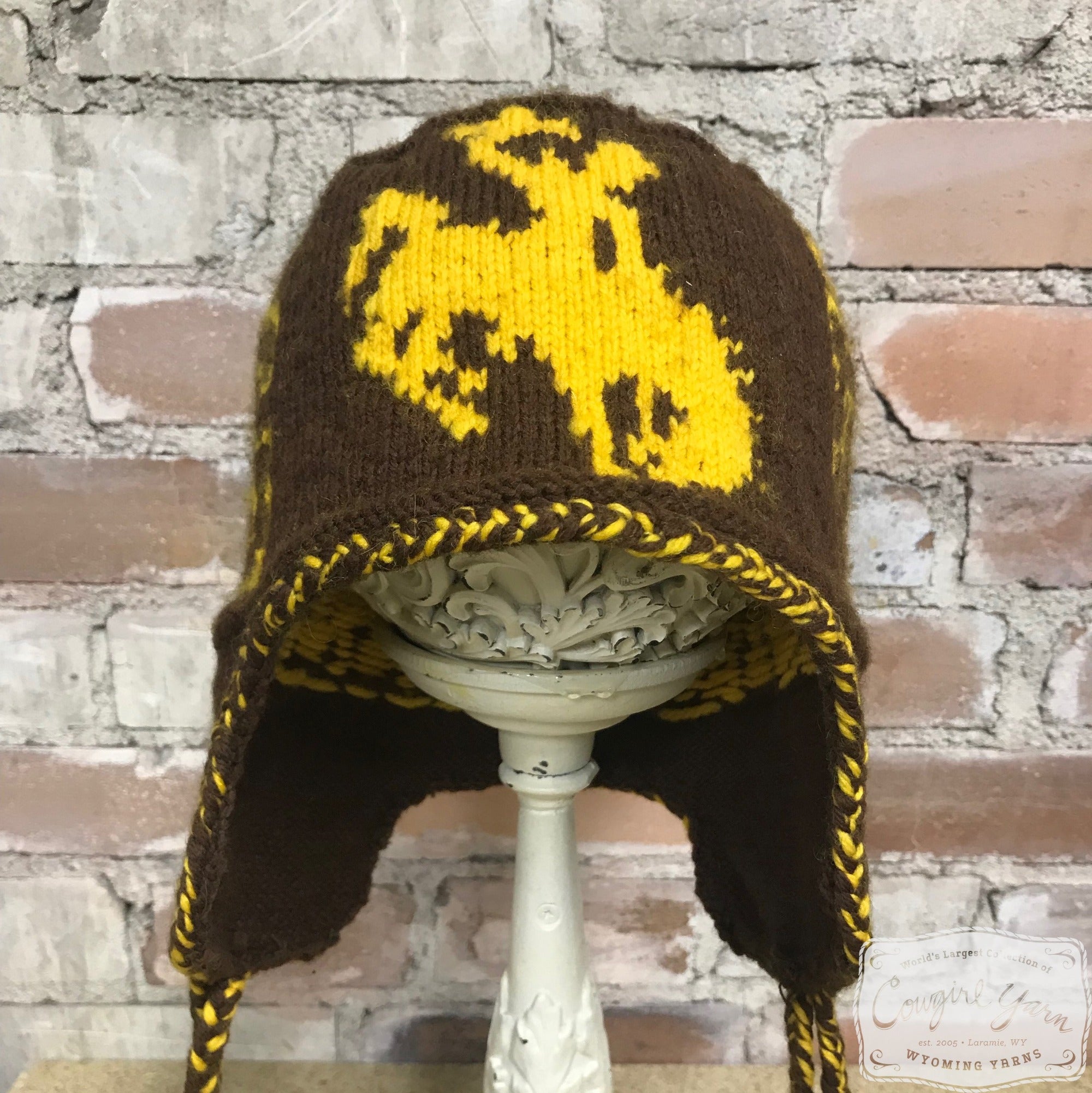 A brown knitted ski hat with a gold colored Steamboat icon against a brick wall.