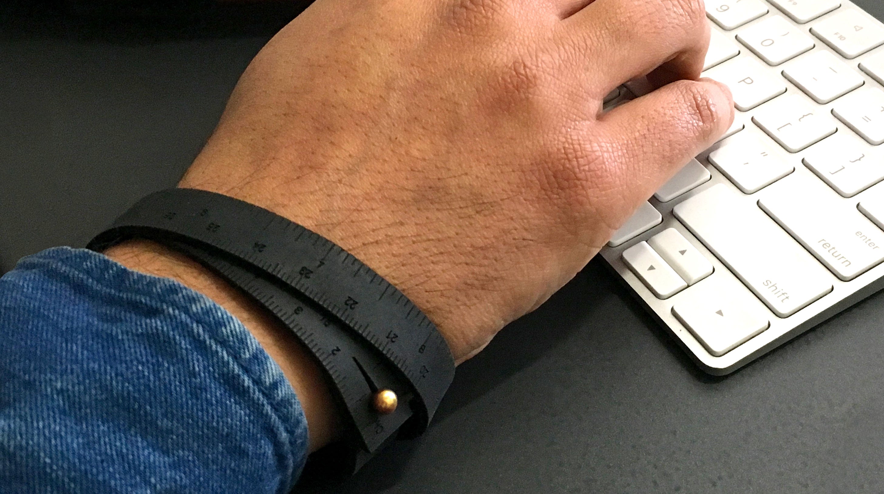A photo of a black wrist ruler with engraved measurements on a man's wrist at the computer keyboard